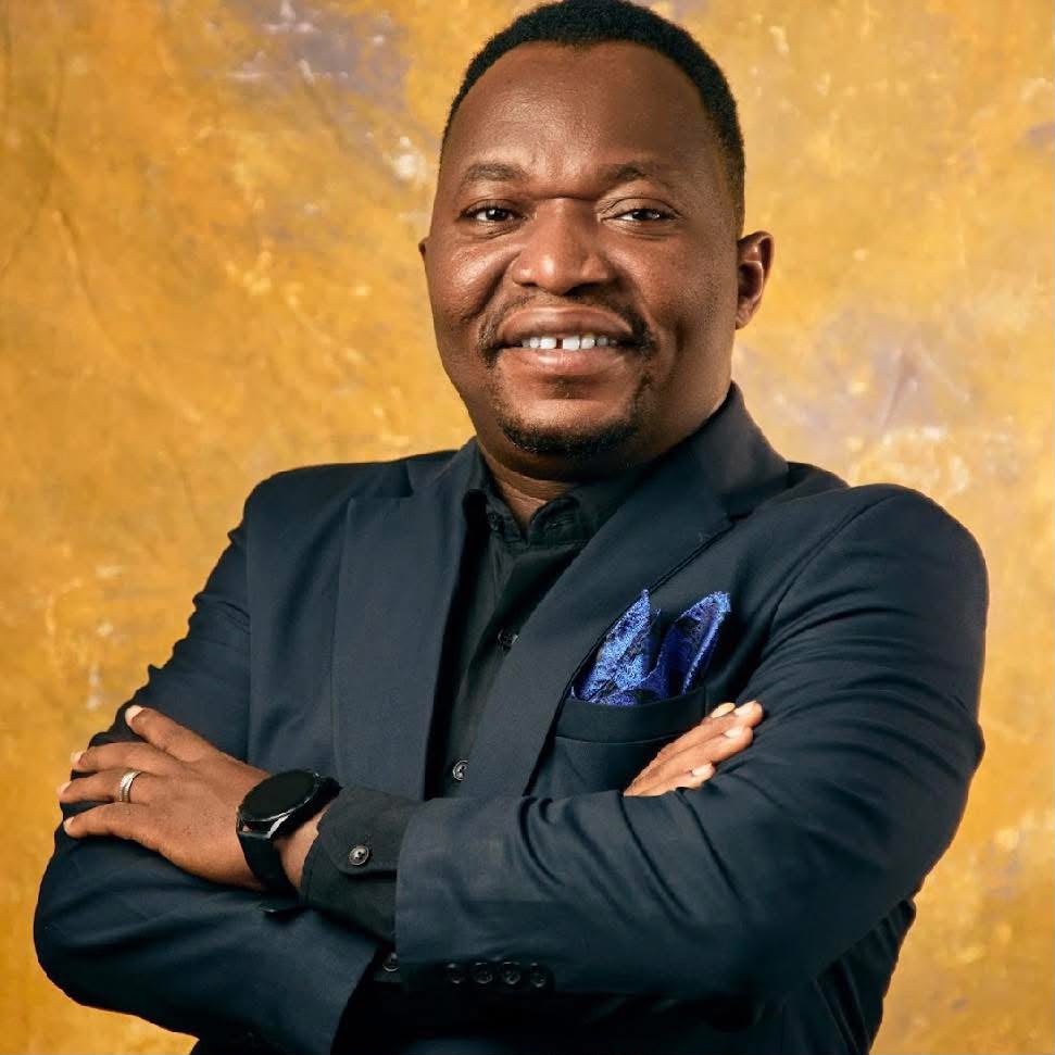 Pastor Ifeanyi Uwakwe in a navy suit with a blue pocket square, arms folded, before a gold backdrop.