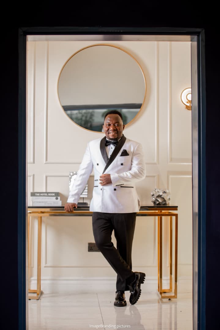 Pastor Ifeanyi Uwakwe in a white dinner jacket and black bow tie, leaning against a console table beneath a round mirror.