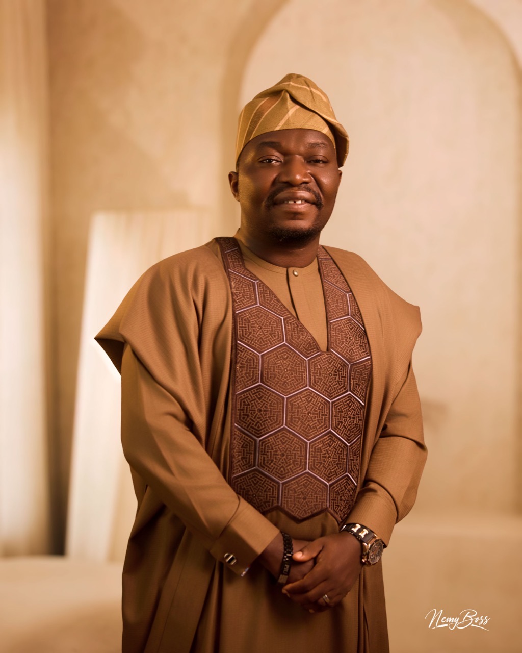 Pastor Ifeanyi Uwakwe in a tan agbada with an embroidered panel and matching cap, hands clasped.