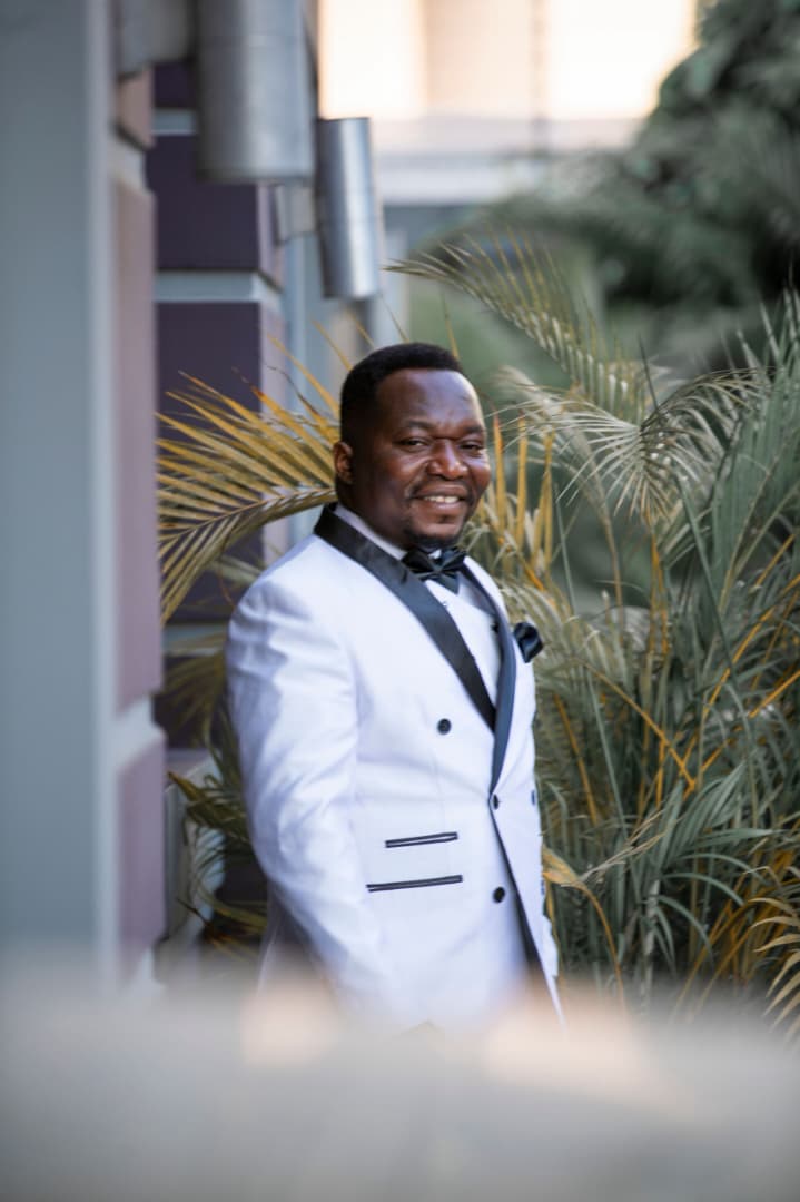 Pastor Ifeanyi Uwakwe in a white dinner jacket outdoors, framed by palm fronds in evening light.