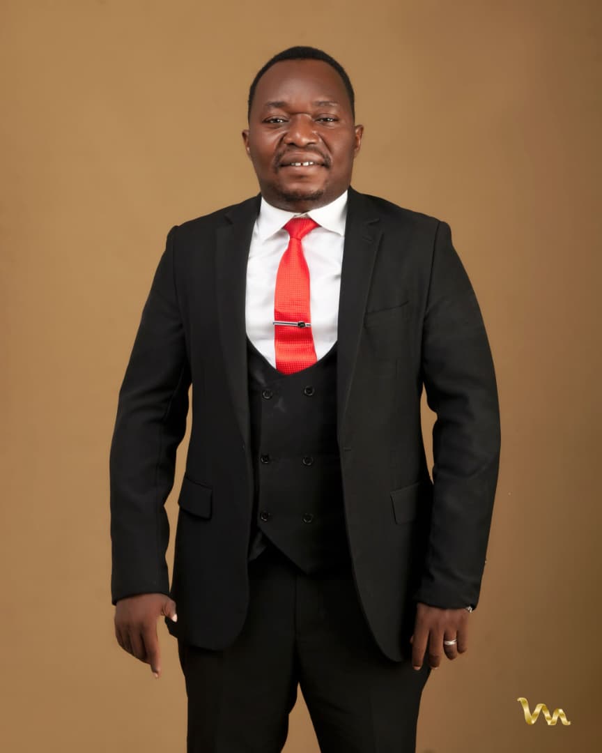 Pastor Ifeanyi Uwakwe in a black three-piece suit with a red tie, against a tan studio backdrop.