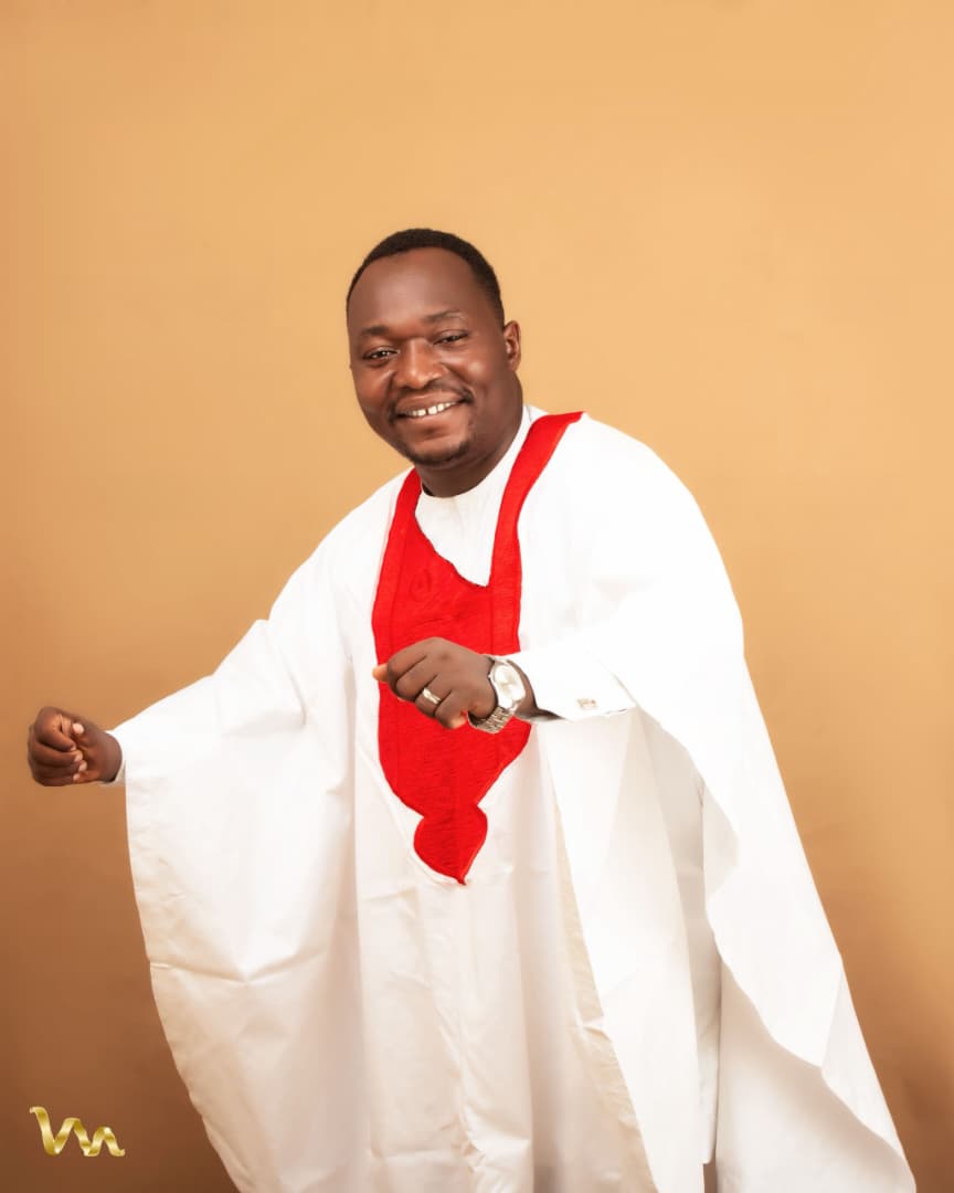 Pastor Ifeanyi Uwakwe smiling in a white agbada with a red embroidered panel, against a warm tan backdrop.