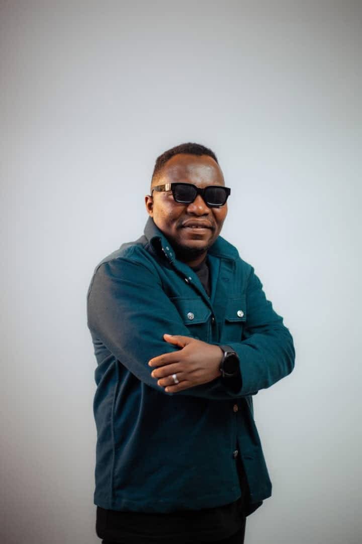 Pastor Ifeanyi Uwakwe in a teal overshirt and dark sunglasses, arms folded against a pale grey background.