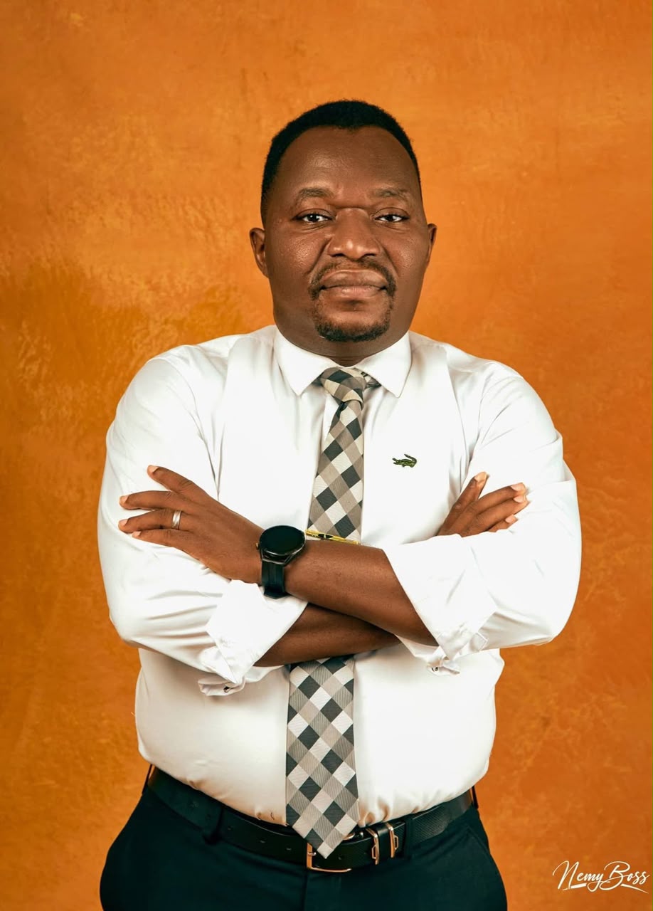 Pastor Ifeanyi Uwakwe in a white shirt and checked tie, arms folded, against a warm ochre backdrop.