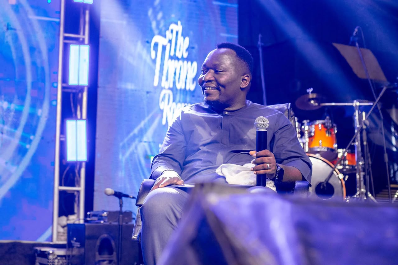 Pastor Ifeanyi Uwakwe seated on stage with a microphone, under blue light at a Throne Room service.